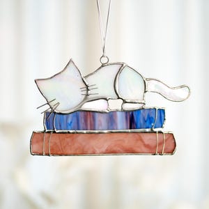 May include: A white stained glass cat figurine is sleeping on top of a stack of stained glass books. The cat has a white iridescent finish and the books are blue and pink.