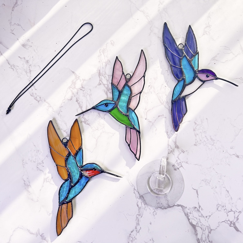 May include: Three stained glass hummingbird ornaments with a black cord and suction cup. The hummingbirds are blue, pink, and orange with green and blue accents.