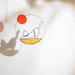 May include: A white cat with a black tail and black ears is depicted in a stained glass circle. The cat is in a yoga pose with its back arched and its head turned to the side. The circle is surrounded by a yellow border and a red circle.