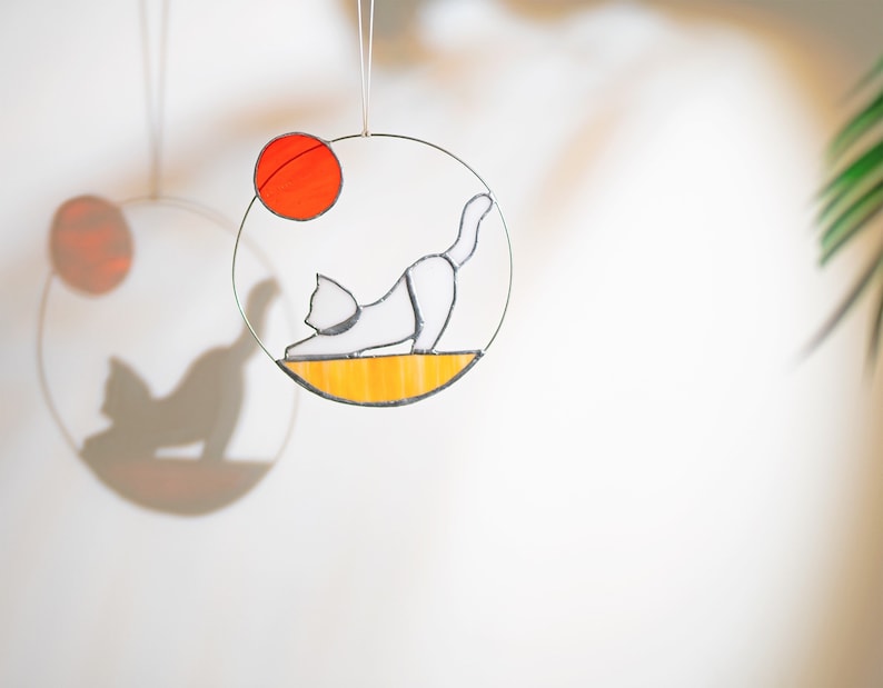 May include: A white cat with a black tail and black ears is depicted in a stained glass circle. The cat is in a yoga pose with its back arched and its head turned to the side. The circle is surrounded by a yellow border and a red circle.