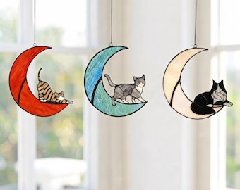 May include: Three stained glass suncatchers shaped like crescent moons, each featuring a cat. The first is a red moon with a brown tabby cat, the second is a blue moon with a grey and white cat, and the third is a cream moon with a black and white cat.