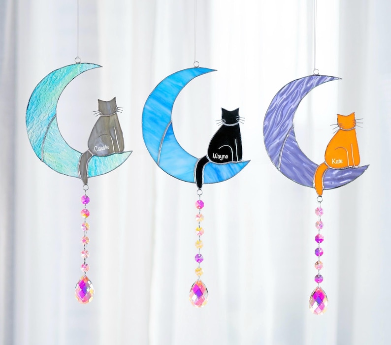 May include: Three stained glass moon and cat suncatchers with crystal beads hanging from the bottom. The moons are blue, purple, and green. The cats are gray, black, and orange. The cats' names are Cassie, Wayne, and Kitie.
