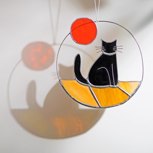 May include: A stained glass suncatcher featuring a black cat sitting on a yellow and orange background with a red circle above the cat.