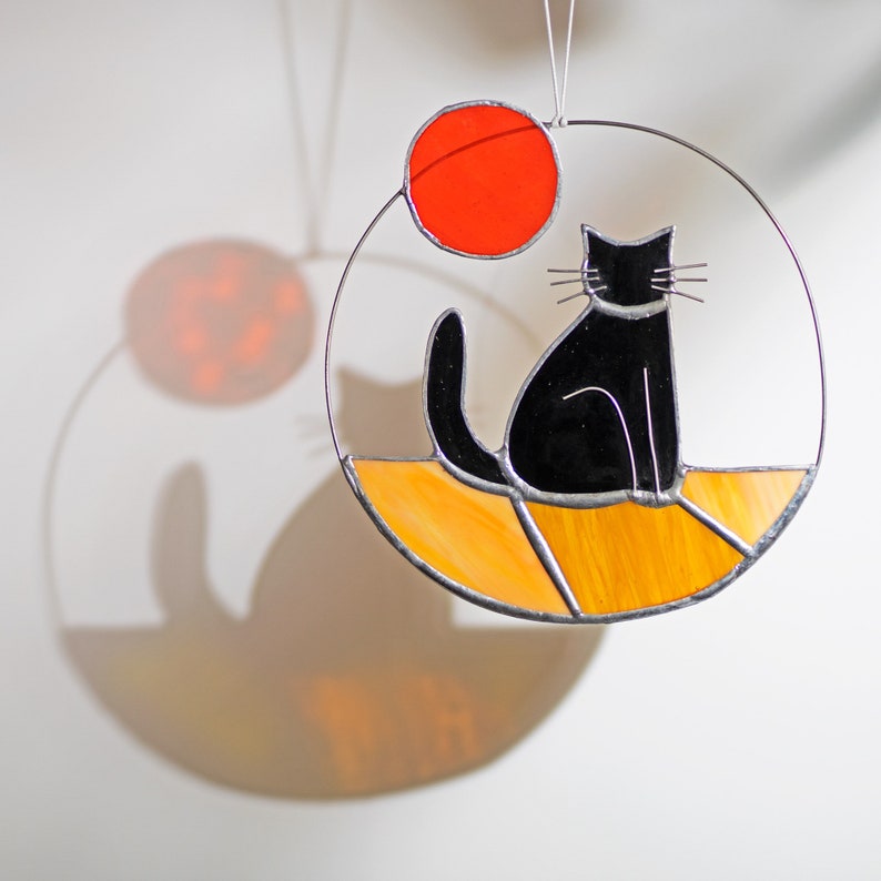 May include: A stained glass suncatcher featuring a black cat sitting on a yellow and orange background with a red circle above the cat.