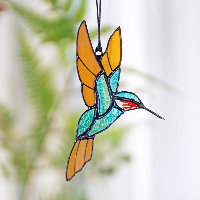 May include: A stained glass hummingbird ornament with a turquoise body, orange wings, and a red beak. The ornament is hanging from a black string.