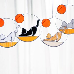 May include: Four stained glass cat suncatchers, each with a different colored cat and a round orange sun. The cats are in a yoga pose.