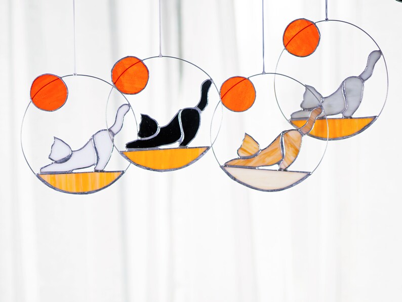 May include: Four stained glass cat suncatchers, each with a different colored cat and a round orange sun. The cats are in a yoga pose.