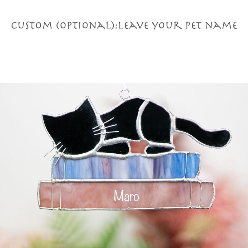 May include: A stained glass ornament of a black cat sleeping on a stack of books. The cat is curled up on the books, with its tail curled around its body. The books are a variety of colors, including blue, purple, and pink. The ornament is personalized with the name "Maro".