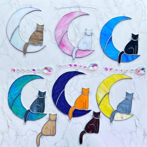 May include: Nine stained glass moon and cat suncatchers. The moons are in various colors, including white, pink, blue, teal, navy, and yellow. The cats are in various colors, including brown, white, black, gray, orange, and tan.