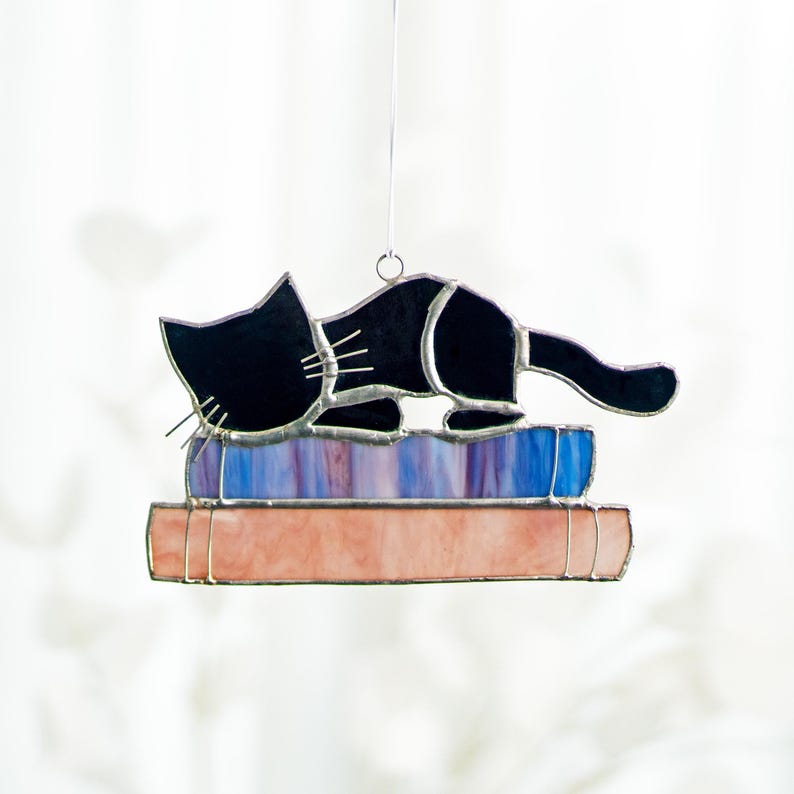 May include: A stained glass ornament depicting a black cat sleeping on top of a stack of books. The books are purple, blue, and pink.