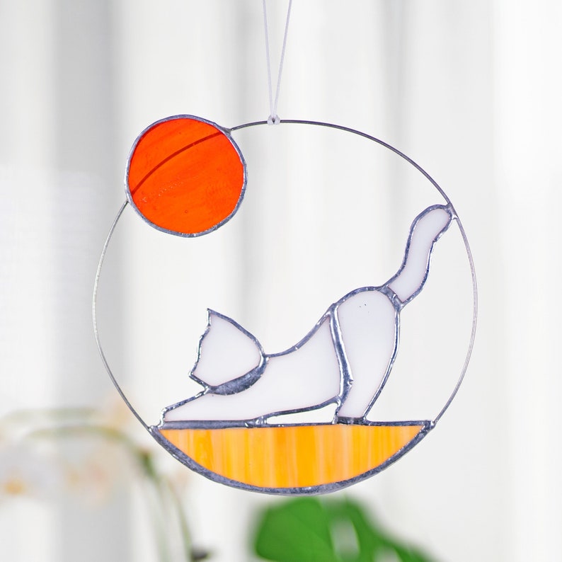 May include: A stained glass suncatcher featuring a white cat in a yoga pose with an orange sun in the background. The cat is on a yellow and orange striped base.