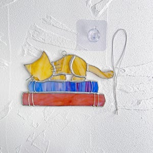 May include: A stained glass cat figurine, with yellow, orange, and white glass, is sleeping on top of a stack of three books. The books are blue, purple, and red. The cat is attached to a string for hanging.