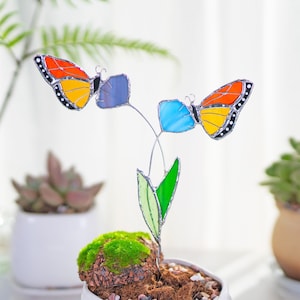 Può includere: Un paletto da giardino in vetro colorato con due farfalle, una blu e una arancione, appollaiate su uno stelo verde con fiori blu e viola.