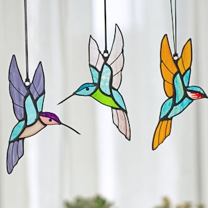 May include: Three stained glass hummingbird ornaments hanging from strings. The hummingbirds are blue, green, and orange with black outlines.
