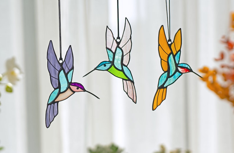 May include: Three stained glass hummingbird ornaments hanging from strings. The hummingbirds are blue, green, and orange with black outlines.