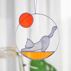 May include: A stained glass sun catcher featuring a white cat in a yoga pose with an orange sun above it. The cat is on a yellow and orange base.