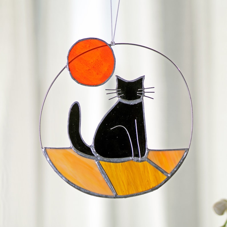 May include: A stained glass suncatcher featuring a black cat sitting on a yellow and orange background with a red circle above the cat.