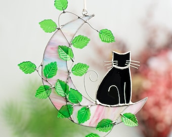 May include: A stained glass moon with a black cat sitting on it. The moon is surrounded by green leaves.