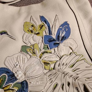 Puede incluir: Bolso de hombro de cuero blanco con bordado floral. El bolso presenta un dise&ntilde;o detallado de flores coloridas, incluyendo flores azules, amarillas y blancas, con detalles de cuentas. Tiene una correa blanca a juego y un cierre plateado en forma de coraz&oacute;n.
