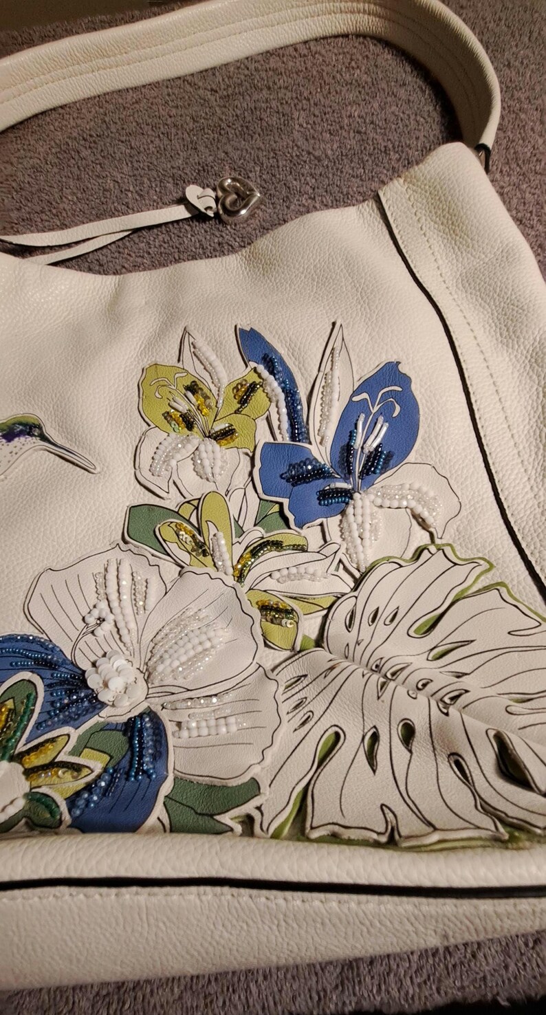 Puede incluir: Bolso de hombro de cuero blanco con bordado floral. El bolso presenta un dise&ntilde;o detallado de flores coloridas, incluyendo flores azules, amarillas y blancas, con detalles de cuentas. Tiene una correa blanca a juego y un cierre plateado en forma de coraz&oacute;n.
