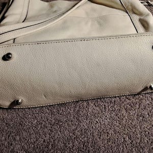 Puede incluir: Bolso de mano de cuero blanco roto con base redondeada y pies plateados. El bolso tiene una correa para el hombro y una superficie texturizada. Las costuras son visibles en los bordes.