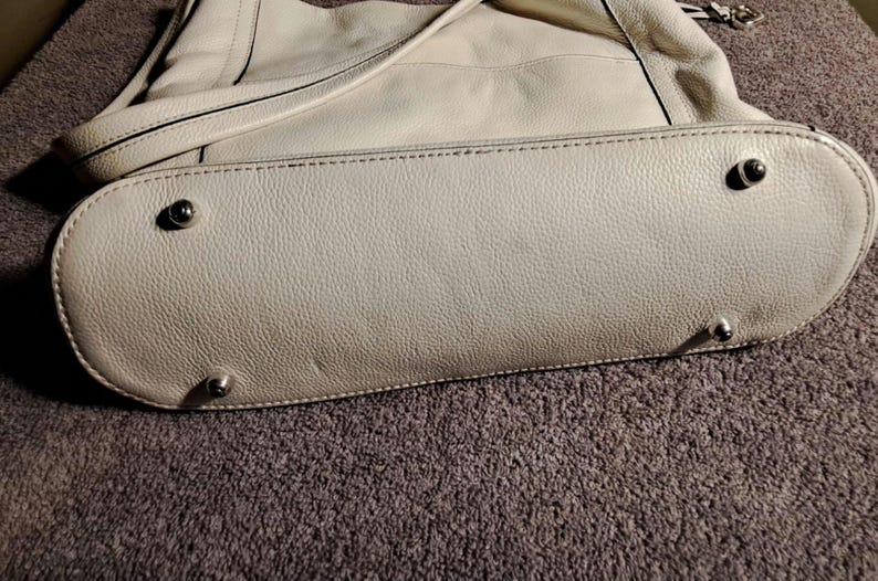 Puede incluir: Bolso de mano de cuero blanco roto con base redondeada y pies plateados. El bolso tiene una correa para el hombro y una superficie texturizada. Las costuras son visibles en los bordes.