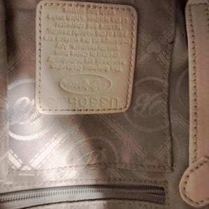 Puede incluir: Primer plano del interior de un bolso de cuero color crema. Se ve una etiqueta rectangular de cuero con el logotipo y el texto de Brighton Collection. El forro del bolso tiene un patr&oacute;n sutil y una cremallera en la parte inferior.