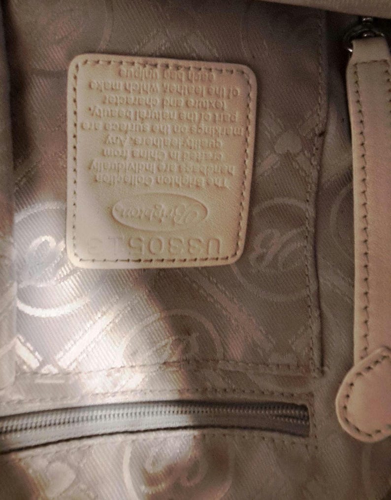 Puede incluir: Primer plano del interior de un bolso de cuero color crema. Se ve una etiqueta rectangular de cuero con el logotipo y el texto de Brighton Collection. El forro del bolso tiene un patr&oacute;n sutil y una cremallera en la parte inferior.