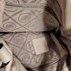 Puede incluir: Bolso de mano de cuero color crema con exterior texturizado. El interior presenta un forro gris con un patr&oacute;n repetido y un bolsillo con cremallera. Una peque&ntilde;a etiqueta rectangular es visible dentro del bolso.