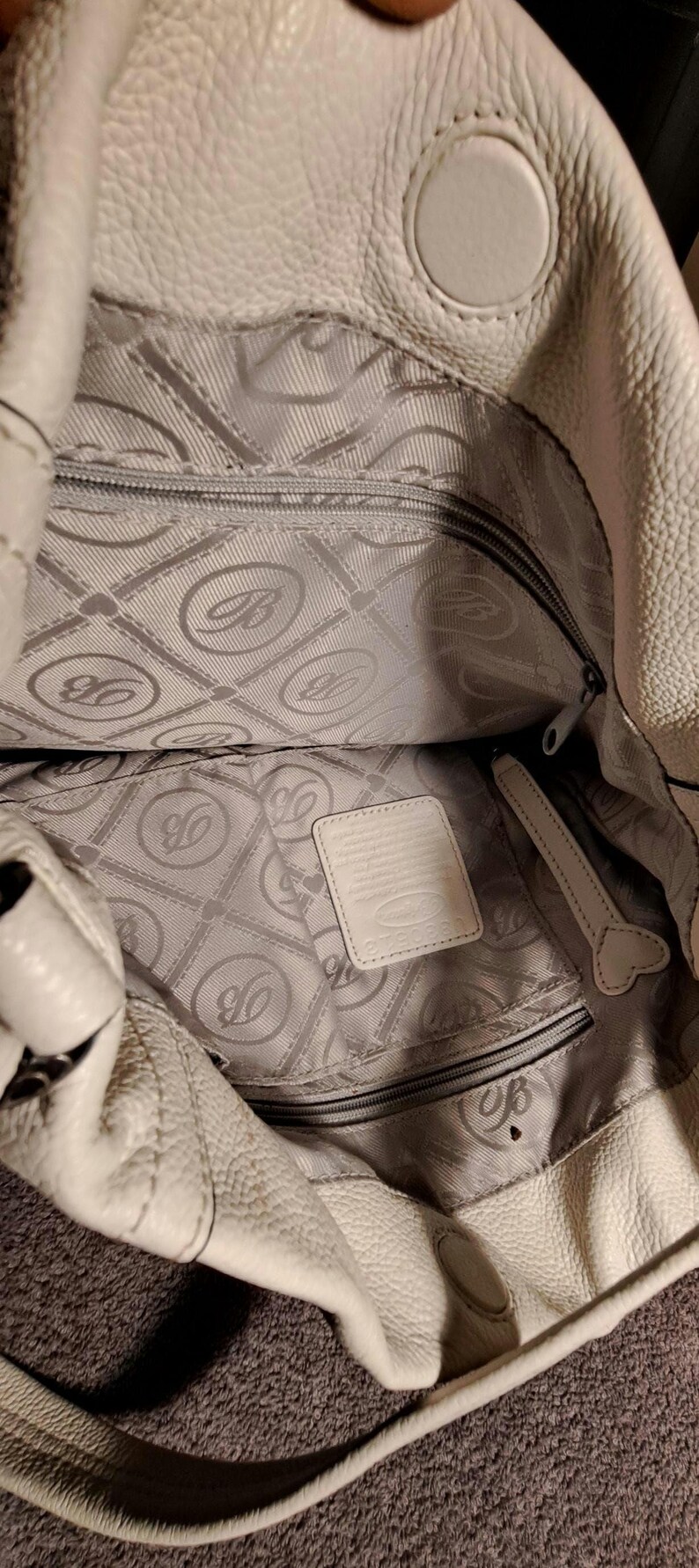 Puede incluir: Bolso de mano de cuero color crema con exterior texturizado. El interior presenta un forro gris con un patr&oacute;n repetido y un bolsillo con cremallera. Una peque&ntilde;a etiqueta rectangular es visible dentro del bolso.