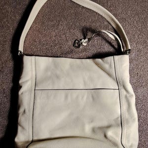 Puede incluir: Bolso de hombro de cuero color crema con una correa larga. El bolso tiene forma rectangular con una costura horizontal en la parte delantera y ribetes negros. Un dije de metal plateado cuelga de la correa.