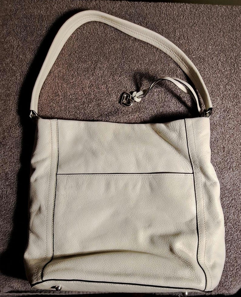 Puede incluir: Bolso de hombro de cuero color crema con una correa larga. El bolso tiene forma rectangular con una costura horizontal en la parte delantera y ribetes negros. Un dije de metal plateado cuelga de la correa.