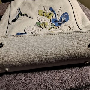 Puede incluir: Un bolso gris claro con un dise&ntilde;o floral y un colibr&iacute;. El bolso presenta una superficie texturizada y patas de metal en la parte inferior. Las flores son en tonos azules, amarillos y blancos.