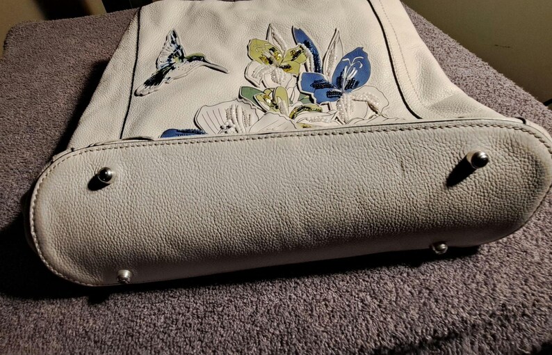 Puede incluir: Un bolso gris claro con un dise&ntilde;o floral y un colibr&iacute;. El bolso presenta una superficie texturizada y patas de metal en la parte inferior. Las flores son en tonos azules, amarillos y blancos.