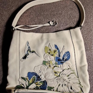 Puede incluir: Bolso de hombro de cuero color crema con un dise&ntilde;o de aplique de colibr&iacute; y flores. El bolso tiene una sola correa para el hombro y un dije en forma de coraz&oacute;n. El dise&ntilde;o floral incluye flores y hojas azules, verdes y blancas.