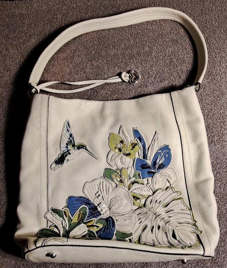 Puede incluir: Bolso de hombro de cuero color crema con un dise&ntilde;o de aplique de colibr&iacute; y flores. El bolso tiene una sola correa para el hombro y un dije en forma de coraz&oacute;n. El dise&ntilde;o floral incluye flores y hojas azules, verdes y blancas.