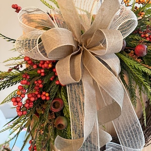 May include: A fall-themed wreath with a large burlap bow and accents of red berries, green foliage, and small red apples. The wreath is designed for seasonal home decor.