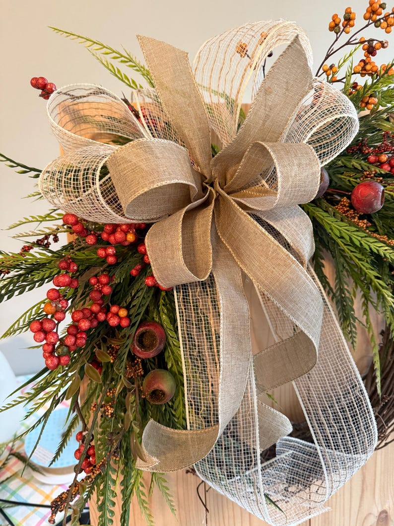 May include: A fall-themed wreath with a large burlap bow and accents of red berries, green foliage, and small red apples. The wreath is designed for seasonal home decor.