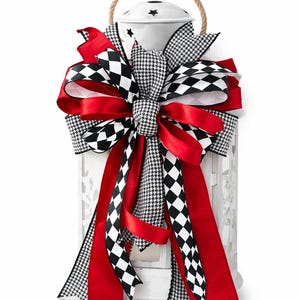 May include: A white metal lantern with a rope handle is decorated with a large bow. The bow features red, black and white ribbons in houndstooth and diamond patterns. The lantern has star cutouts.