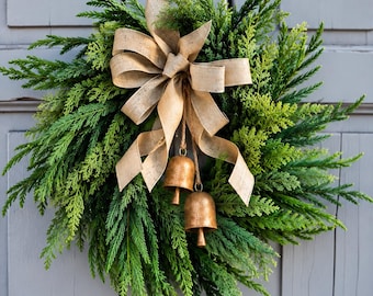 May include: A circular wreath made of vibrant green cedar foliage, featuring a large, centered beige burlap bow. Two metallic, bronze-toned bells hang from the center of the bow by thin twine, resting against the dense, textured evergreen branches.