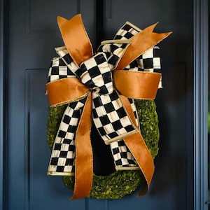 May include: A decorative wreath featuring a moss base, adorned with a large bow. The bow has a black and white checkered pattern with gold trim, and is accented with wide, solid orange ribbons. The wreath is hanging on a dark blue door.