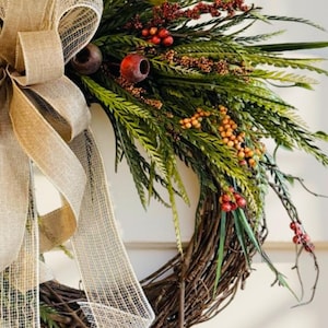 May include: A rustic wreath with a natural twig base, adorned with green foliage, red berries, and orange accents. A large burlap bow adds a touch of elegance. Perfect for seasonal home decor.