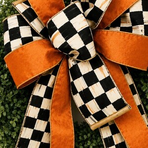 May include: A decorative bow featuring alternating ribbons of solid burnt orange and a black and white checkered pattern. The bow is layered with multiple loops and long, flowing tails, secured in the center, and positioned atop a green wreath.