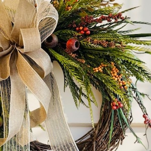 May include: A rustic wreath with a brown twig base, adorned with green foliage, red berries, and orange accents. A large burlap bow with a mesh overlay adds a touch of elegance. Perfect for seasonal home decor.