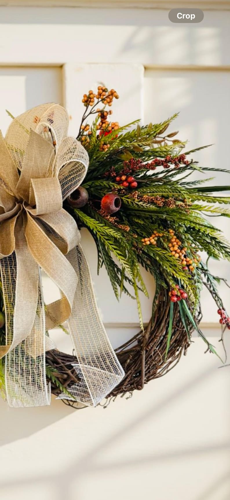 May include: A rustic wreath with a brown twig base, adorned with green foliage, red berries, and orange accents. A large burlap bow with a mesh overlay adds a touch of elegance. Perfect for seasonal home decor.