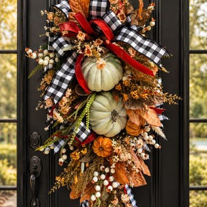 May include: A teardrop-shaped door swag featuring two large sage green pumpkins and one smaller orange pumpkin. The arrangement includes black and white buffalo check ribbon, a red ribbon, dried brown leaves, white berries, and small orange flowers.