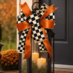 May include: A black metal lantern featuring a large decorative bow on top. The bow is crafted from orange velvet ribbon and black and white checkered ribbon. Inside the lantern are two white pillar candles nestled in green foliage.