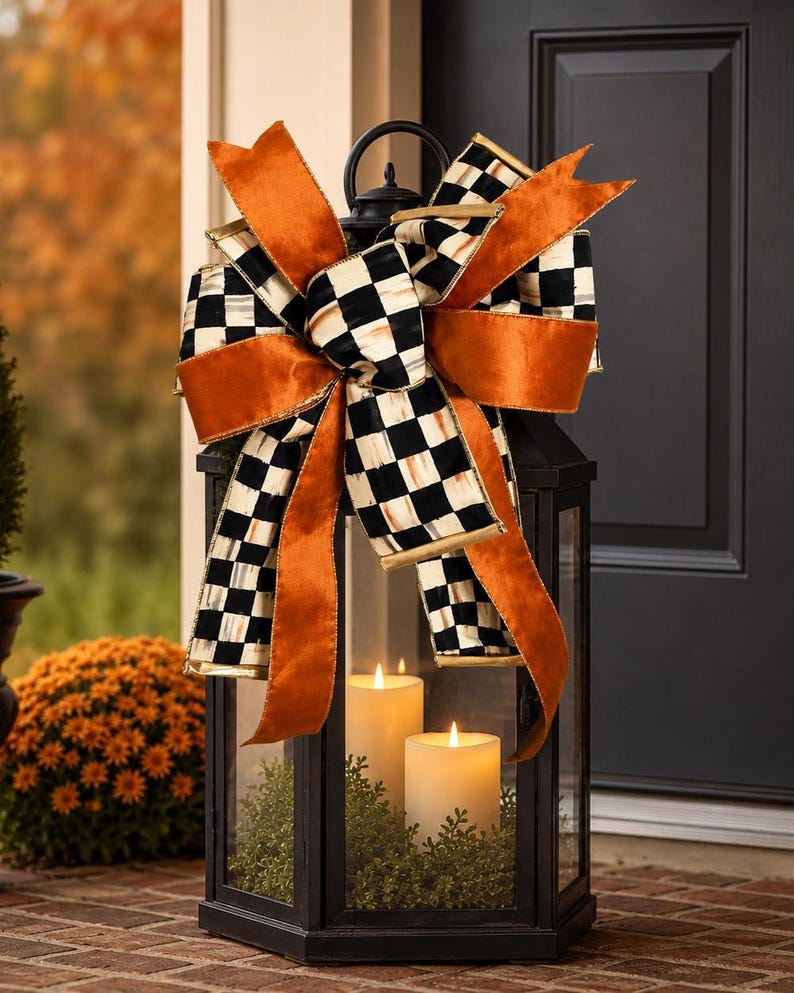 May include: A black metal lantern featuring a large decorative bow on top. The bow is crafted from orange velvet ribbon and black and white checkered ribbon. Inside the lantern are two white pillar candles nestled in green foliage.