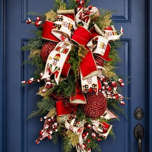 May include: A teardrop-shaped holiday wreath featuring green pine branches, red glittery ornaments, and red berries. The arrangement is accented with wide ribbons featuring a nutcracker pattern, solid red velvet ribbons, and gold glittery foliage.