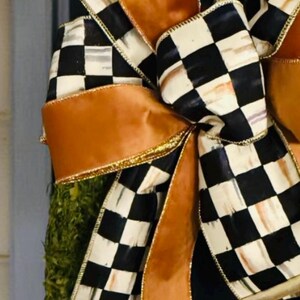 May include: A decorative bow featuring a combination of solid copper-colored ribbon and a black and white checkered pattern ribbon. The bow has multiple loops and long trailing ends with gold-toned edges, attached to a green textured base.